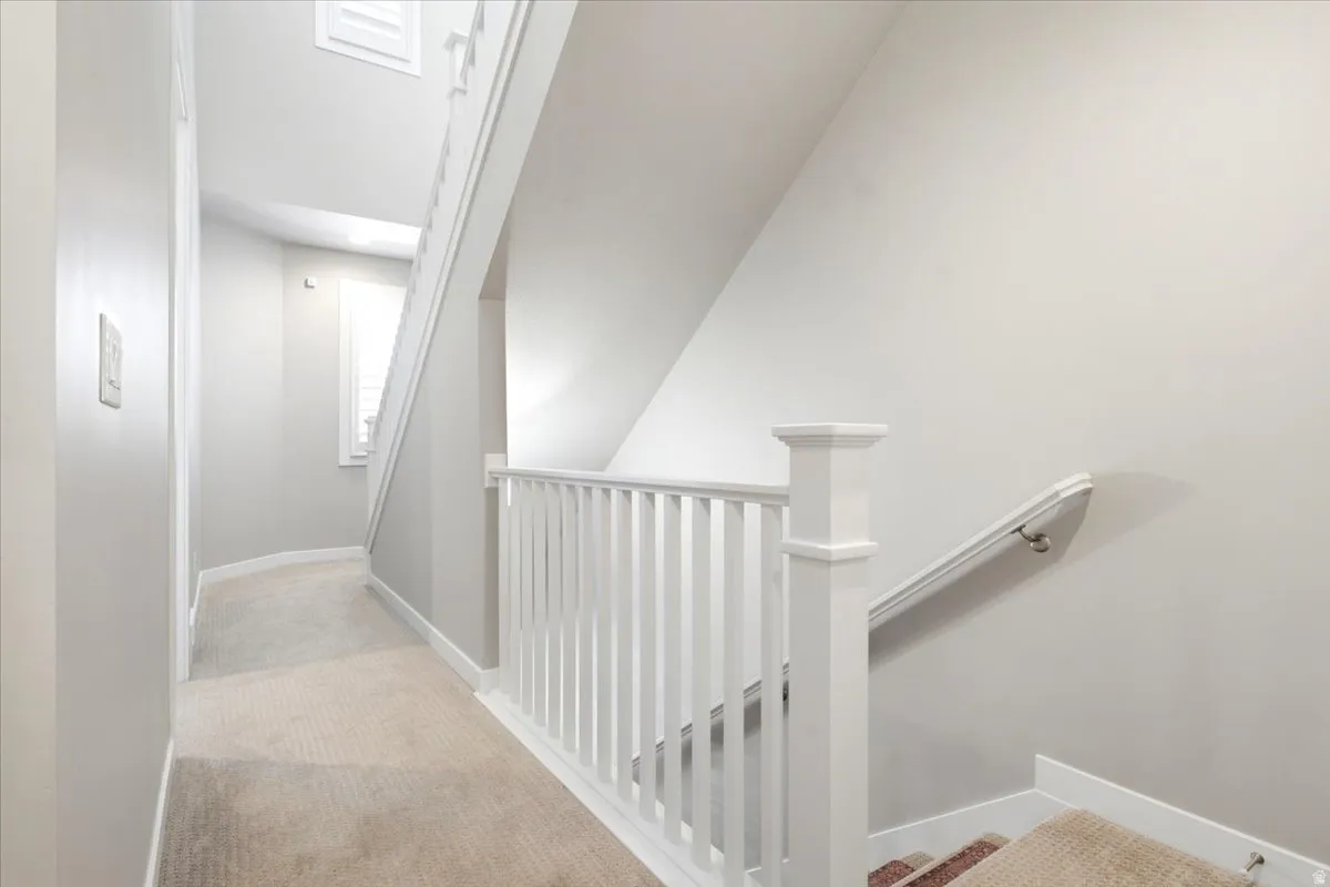 Carpeted hallway with white trim, white spindle railing, and a window with white louvered shutters