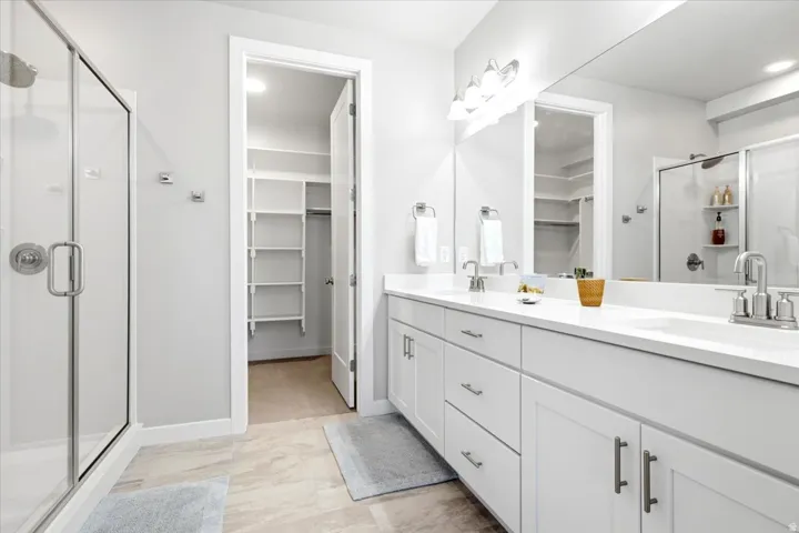 Spacious bathroom featuring a glass-enclosed shower, dual vanities with white countertops, expansive mirrors, and a walk-in closet with built-in shelving