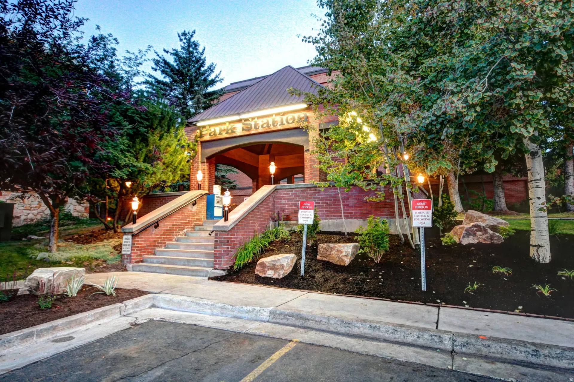 Entryway featuring brick facade, concrete stairs with black railings, a covered portico with overhead lighting, and mature landscaping with trees and decorative rocks