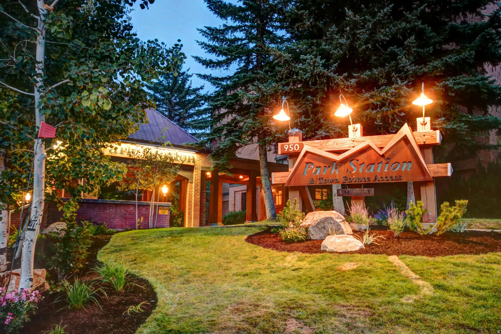 Wood-toned architectural signage with integrated lighting, surrounded by landscaped grounds featuring natural rock elements, mature trees, manicured lawns, and planted flower beds