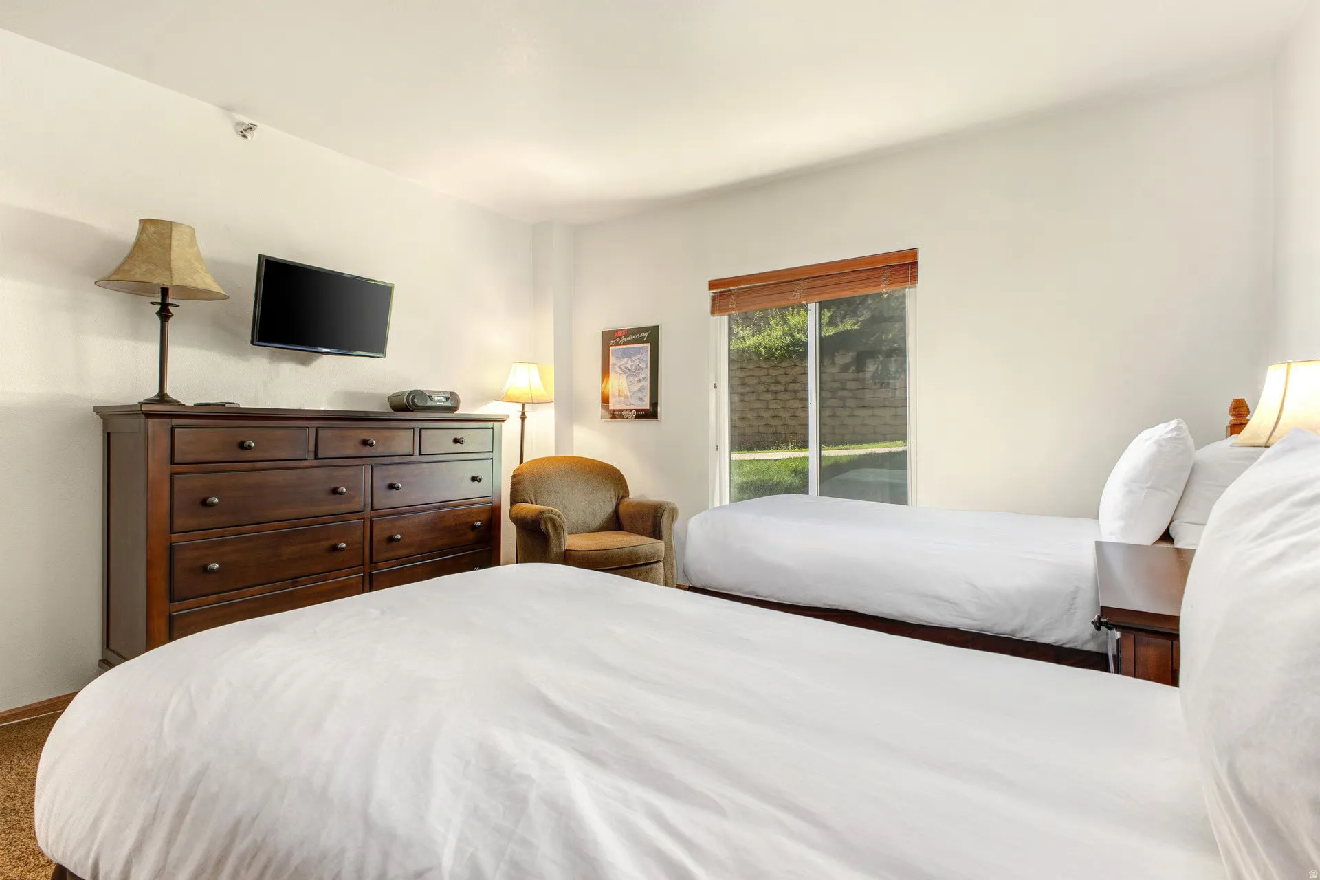 Bedroom featuring two twin-sized beds, a large window with wood blinds, and a wall-mounted television above a dark wood dresser