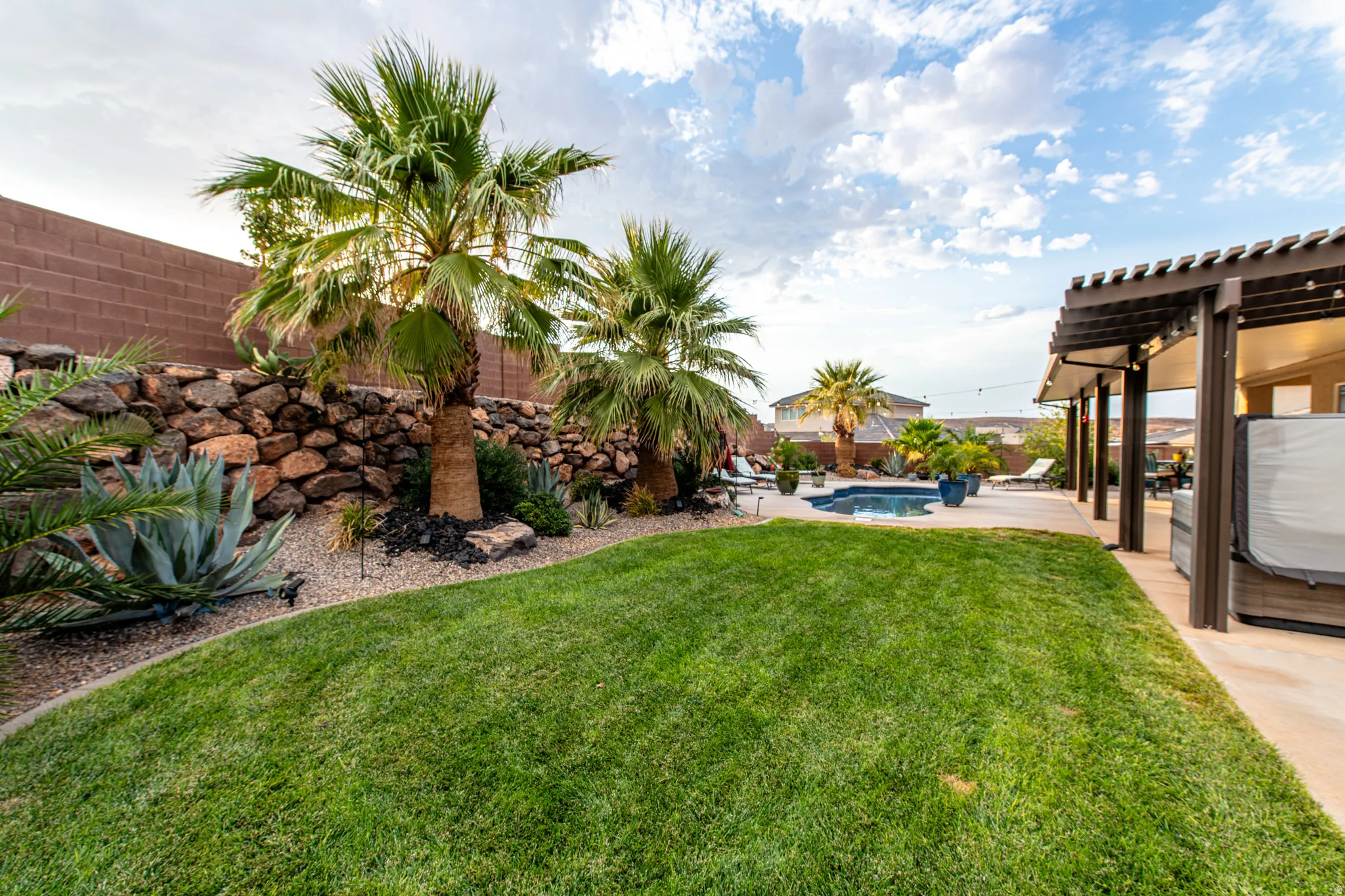 Expansive backyard featuring a lush lawn, rock retaining wall, mature palm trees, and a covered patio with a dark wood pergola