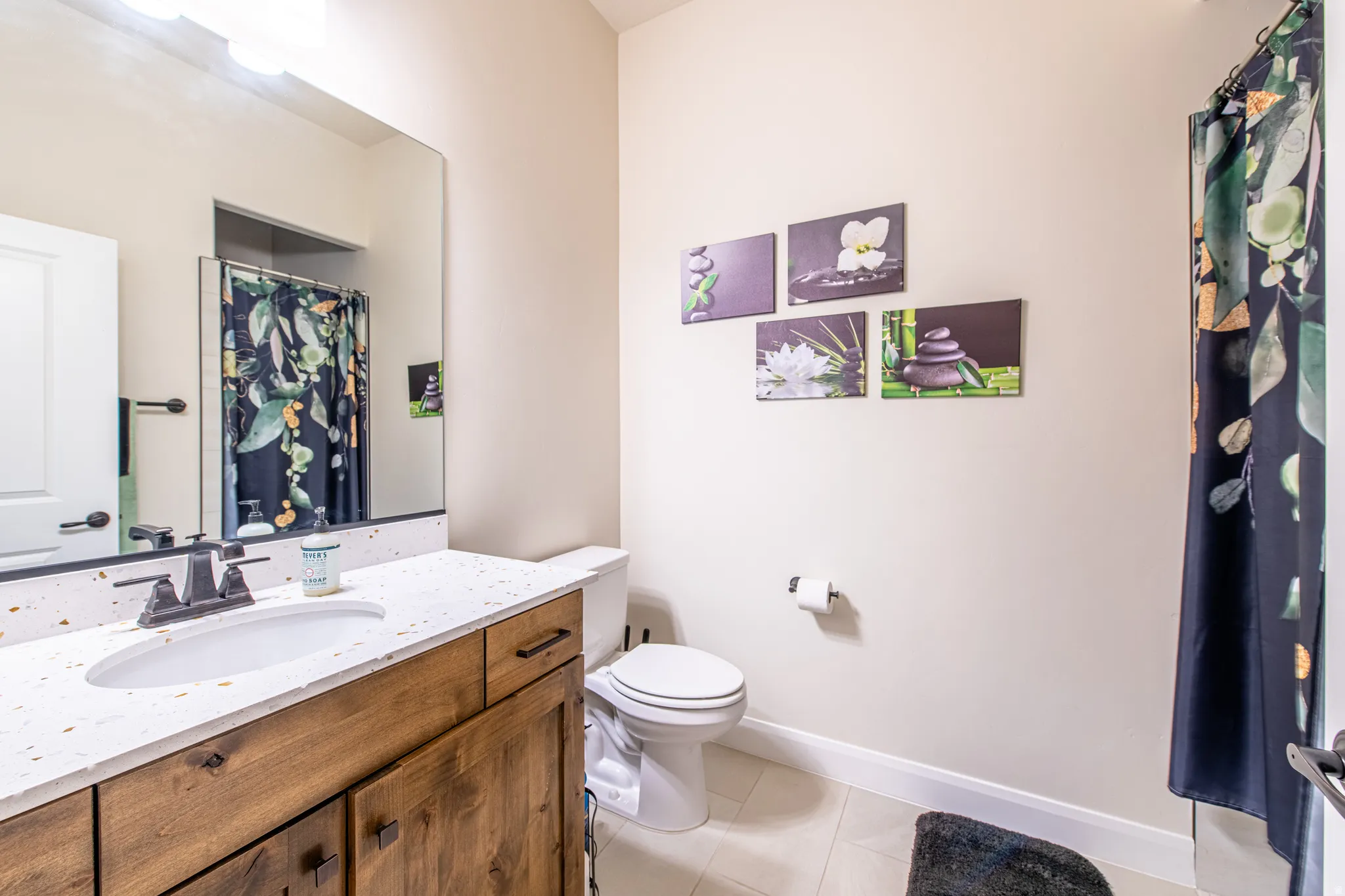 White solid-surface countertop with integrated sink, wood-finish vanity cabinetry with dark hardware, large wall-mounted mirror, and tile flooring