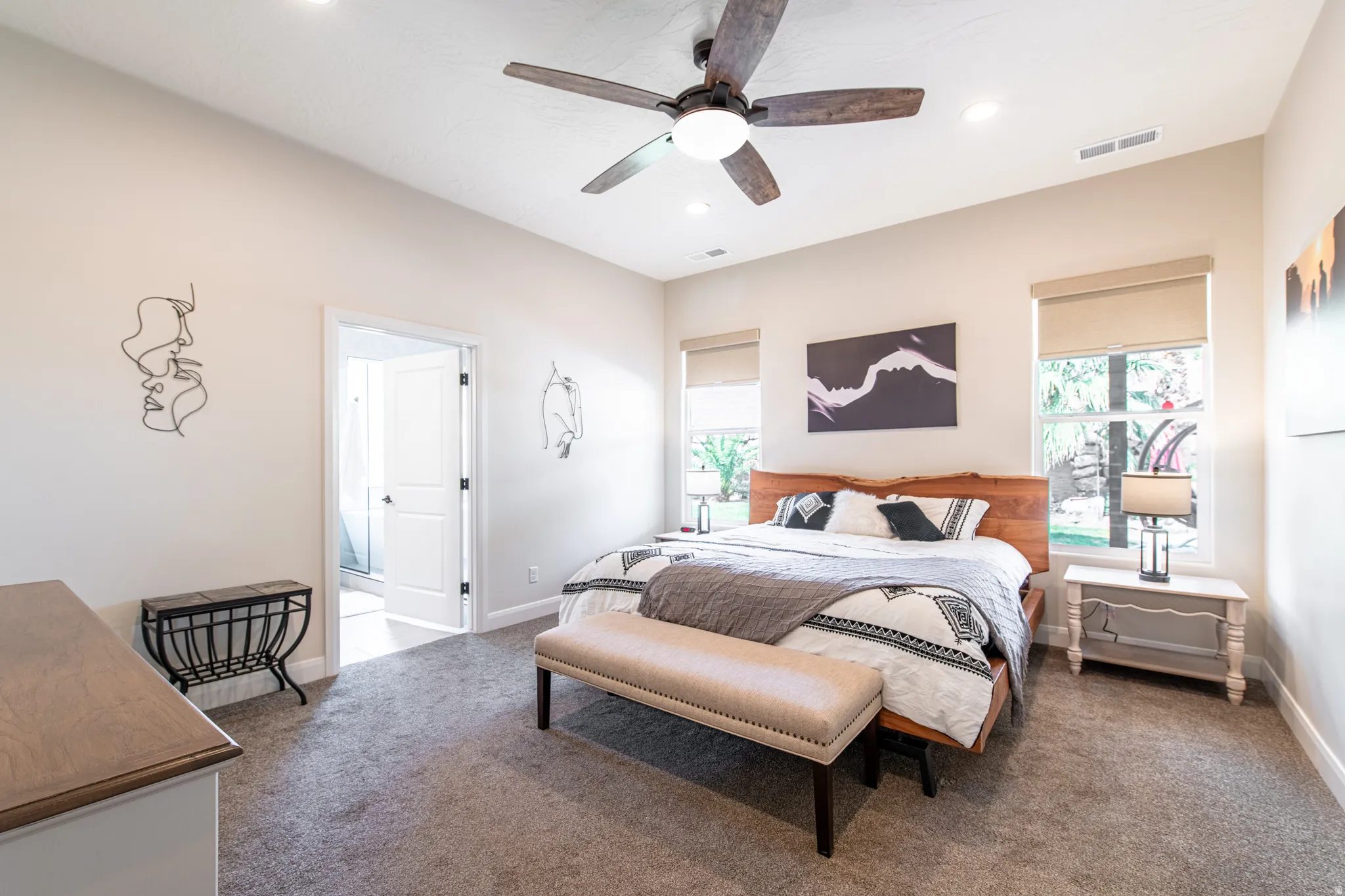 Recessed ceiling lighting, wood-finish ceiling fan, two large windows with roller shades, neutral wall paint, and plush carpet flooring