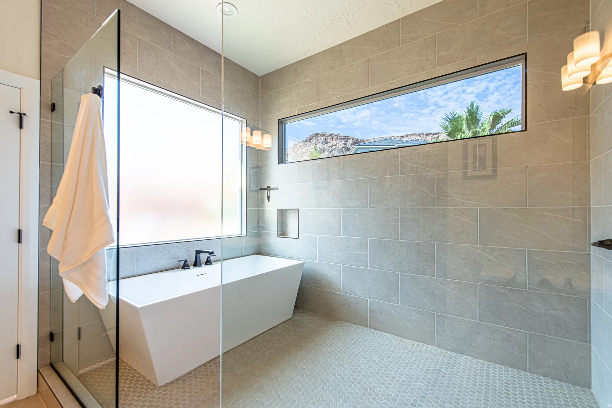 Freestanding soaking tub with matte black fixtures, glass-enclosed shower, light grey rectangular wall tile, hexagonal floor tile, and a rectangular transom window