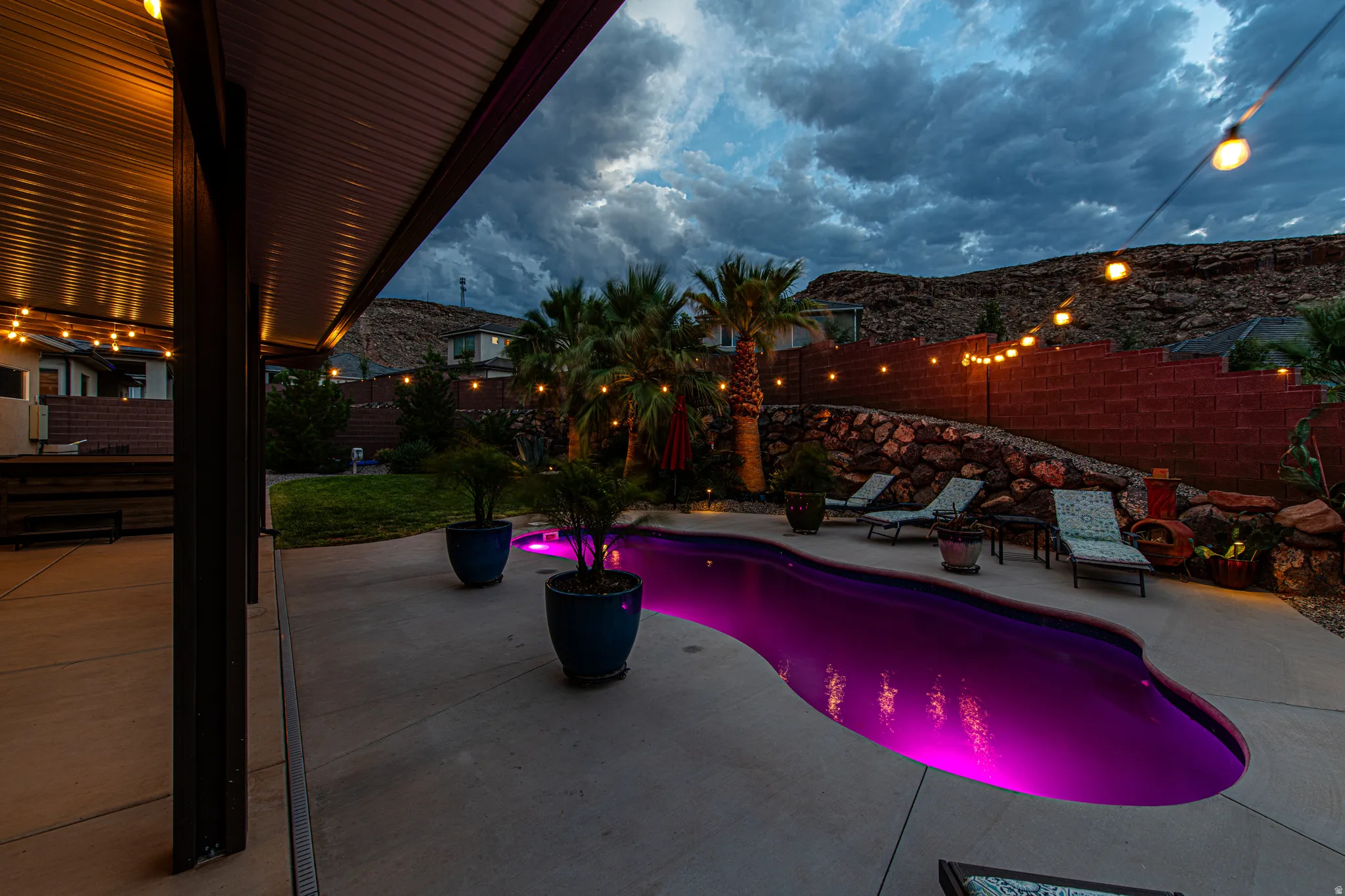 Resort-style backyard featuring a uniquely shaped pool with vibrant lighting, a covered patio, and a terraced rock wall
