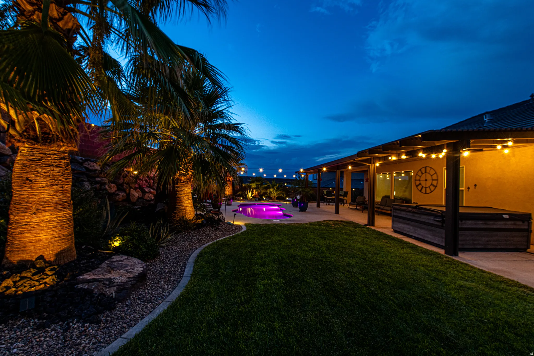 Expansive backyard featuring a manicured lawn, established palm trees with integrated uplighting, a pergola-covered patio with string lighting, a built-in spa, and a swimming pool with integrated lighting