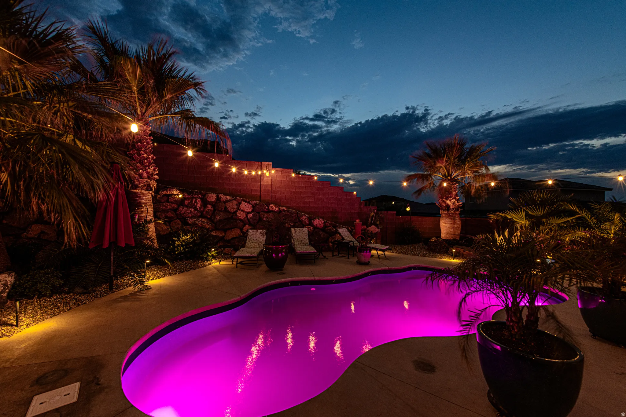 Freeform swimming pool with colored lighting, concrete pool deck, string lighting, mature palm trees, and a tiered rock wall