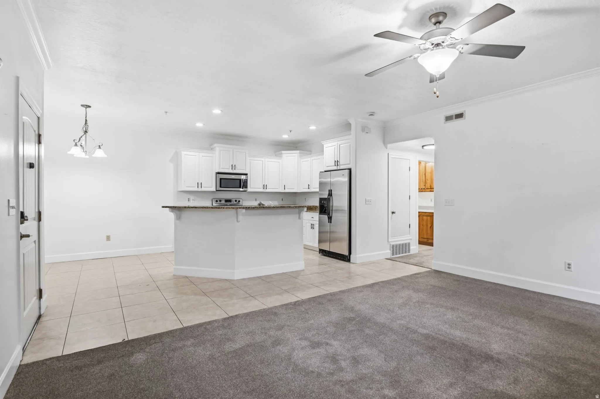 Open-concept kitchen and living area featuring a white cabinetry island with breakfast bar, stainless steel appliances, ceramic tile flooring in the kitchen zone, and plush carpeting in the adjoining living space
