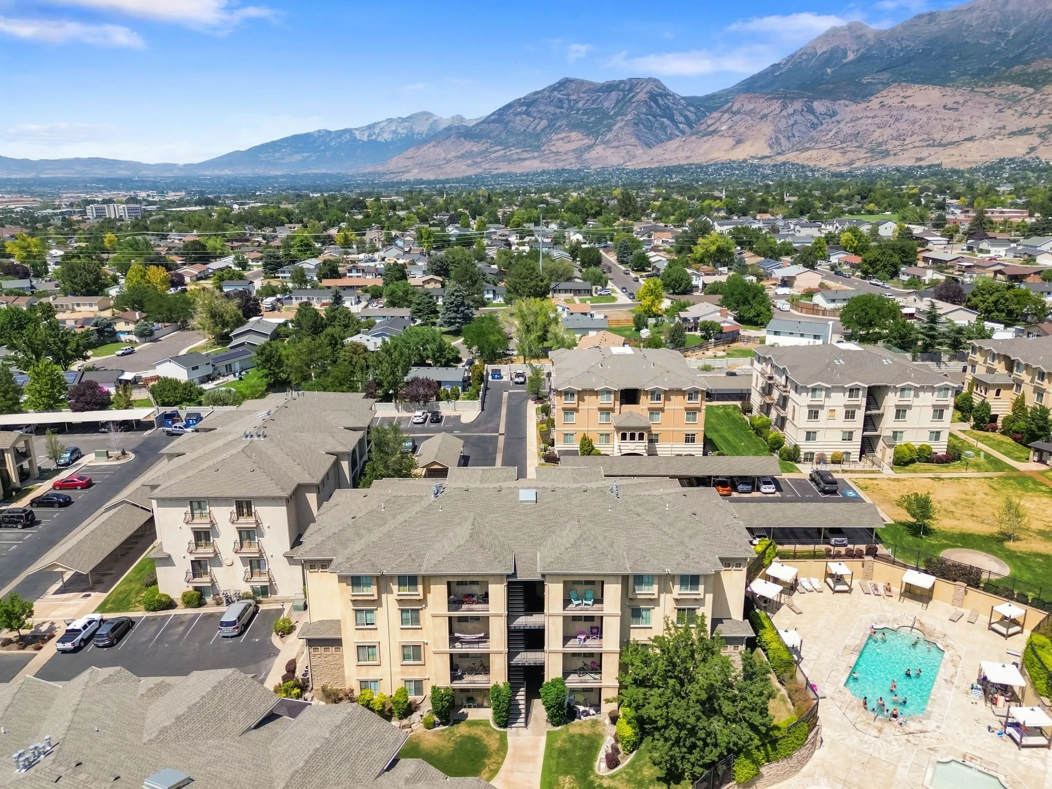 Multi-story buildings with balconies, a swimming pool with surrounding patio, covered parking, and an expansive mountain backdrop