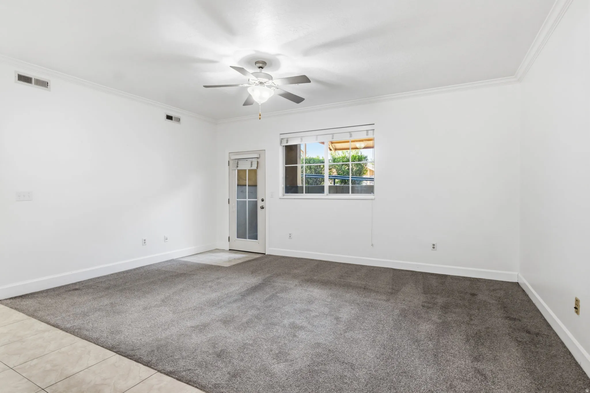 Spacious room featuring neutral white walls, tile and carpeted flooring, ceiling fan with integrated lighting, and glass door opening to natural light