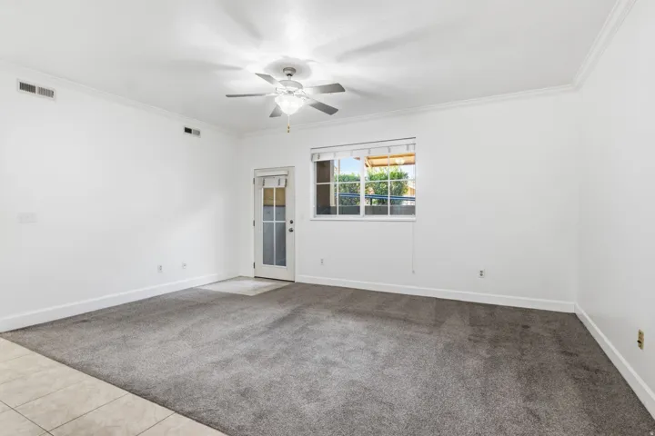 Spacious room featuring neutral white walls, tile and carpeted flooring, ceiling fan with integrated lighting, and glass door opening to natural light