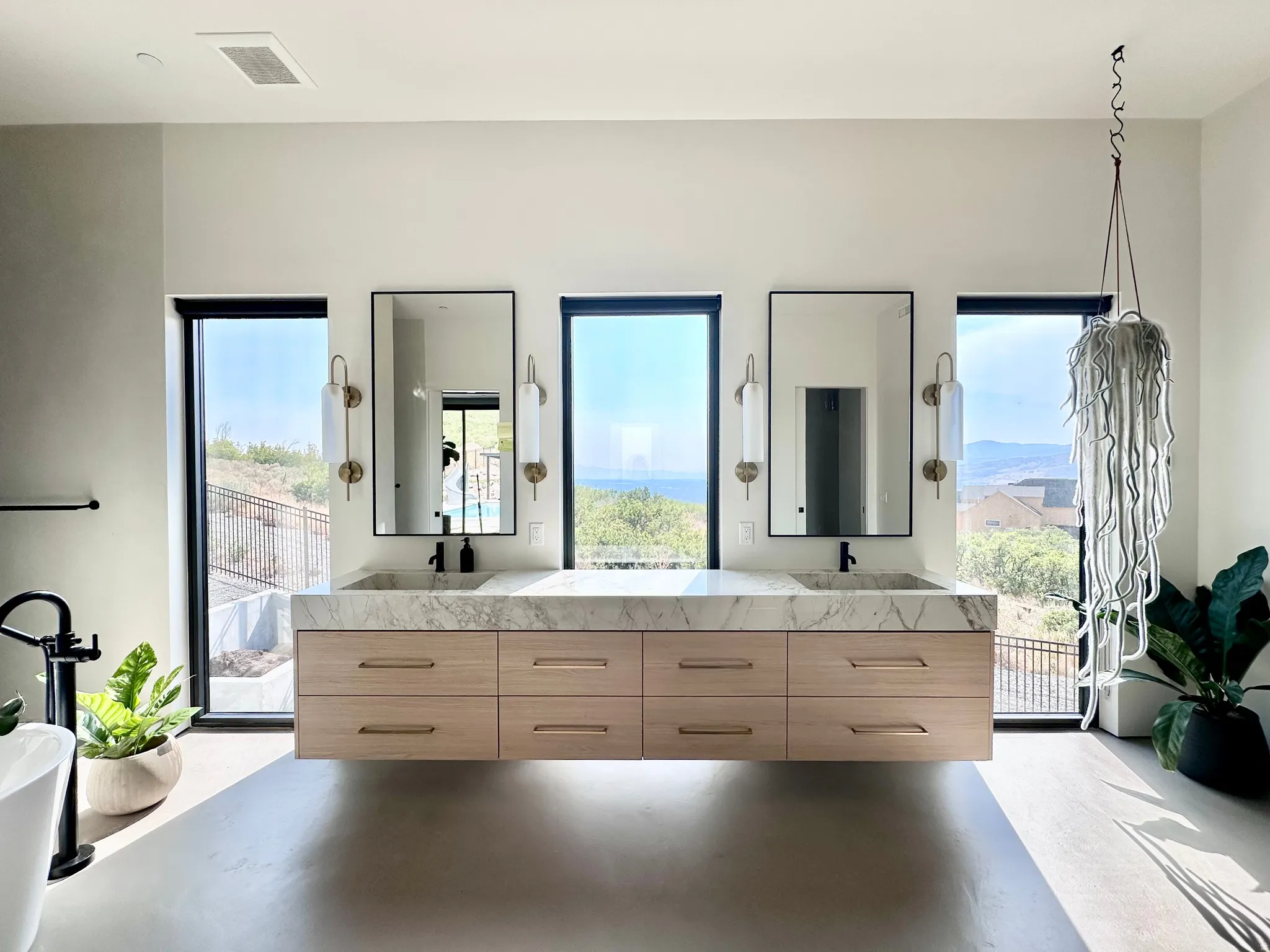 Floating dual vanity with a stone countertop and integrated sinks, flanked by tall framed windows, and illuminated by decorative sconces