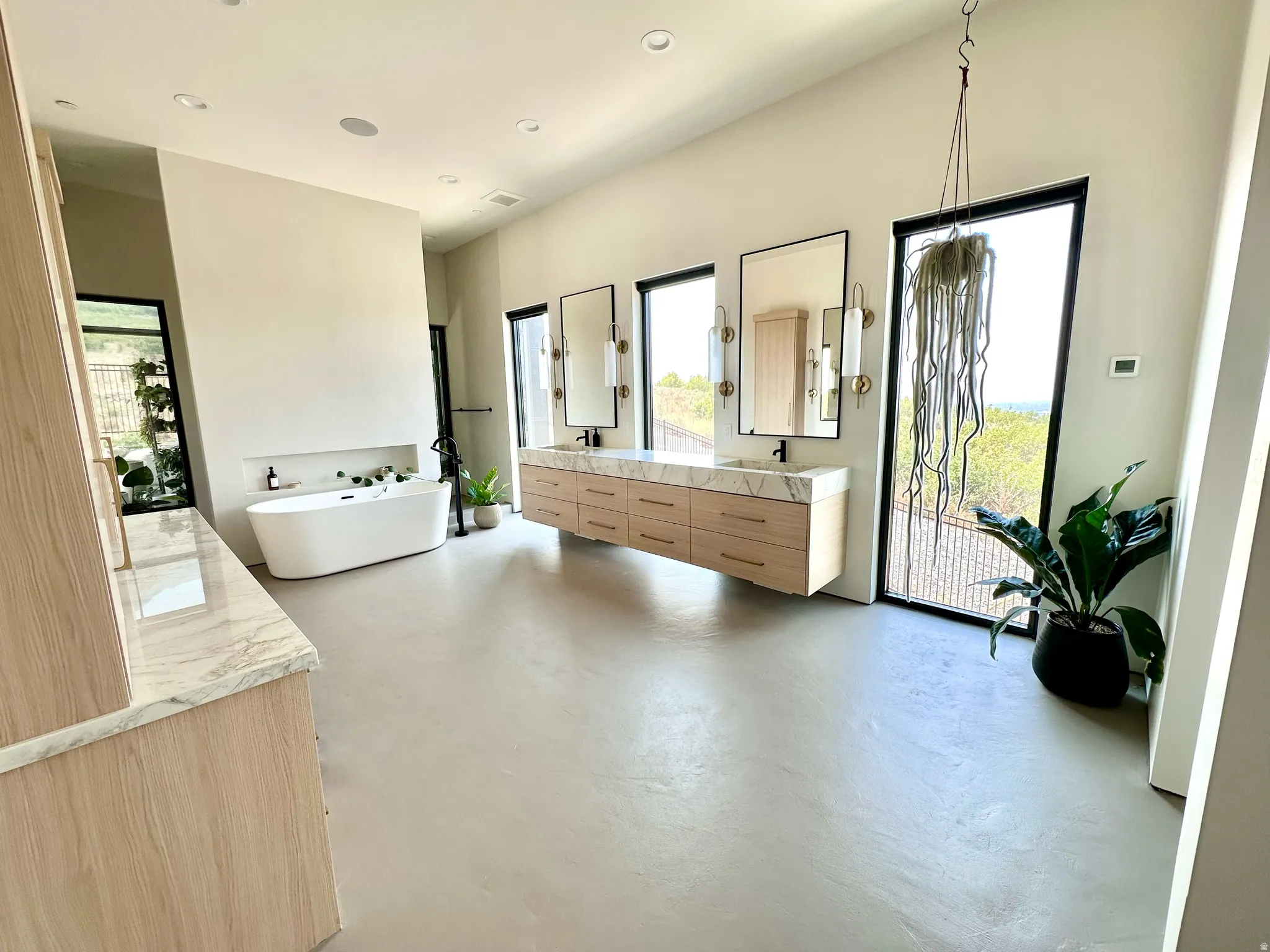 Freestanding soaking tub with a recessed shelf, dual vanity with wood-finish cabinetry and stone countertops, and large framed windows