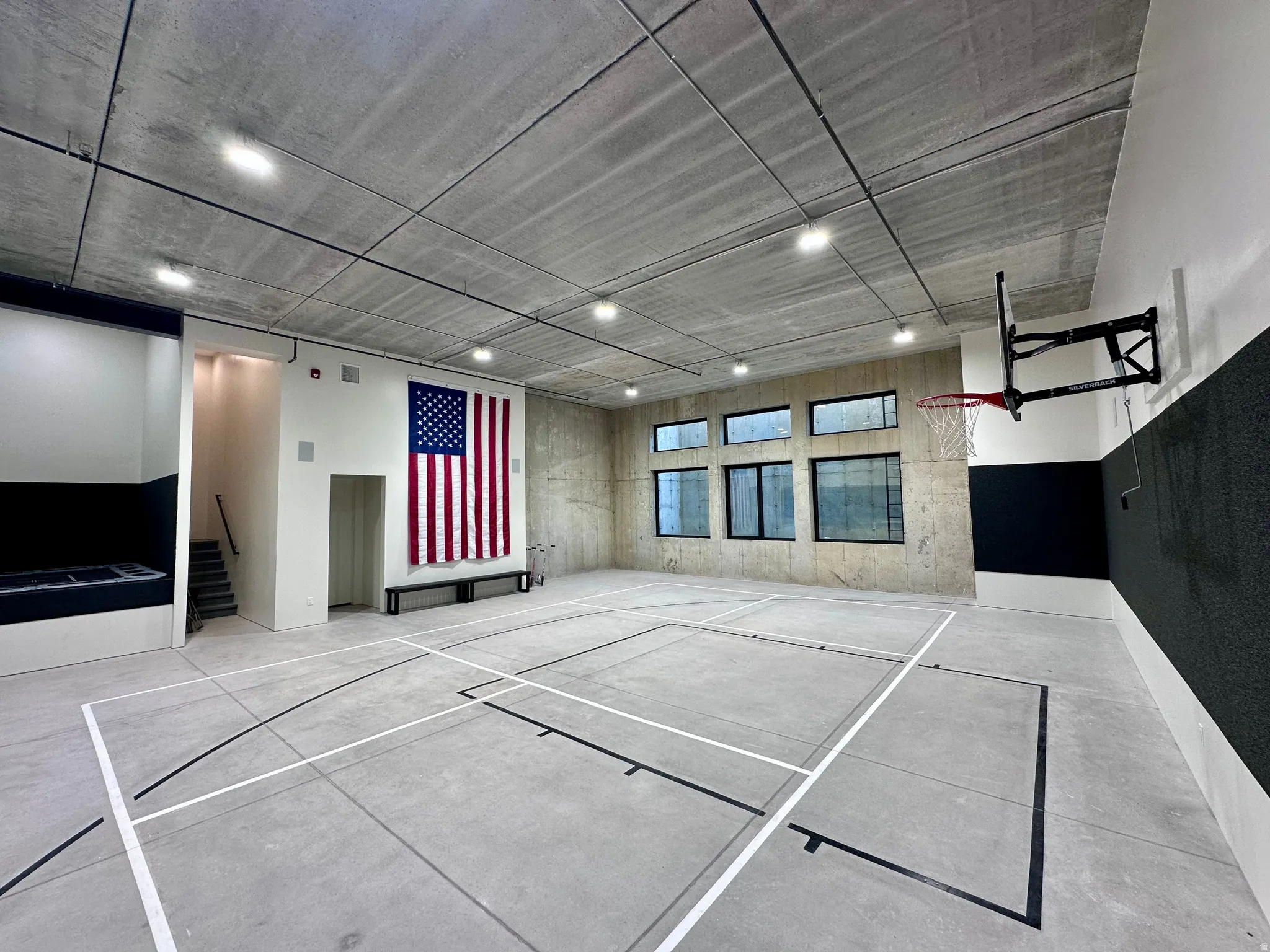 Indoor court featuring concrete flooring with court markings, exposed concrete ceiling with recessed lighting, multiple windows, and a wall-mounted basketball hoop