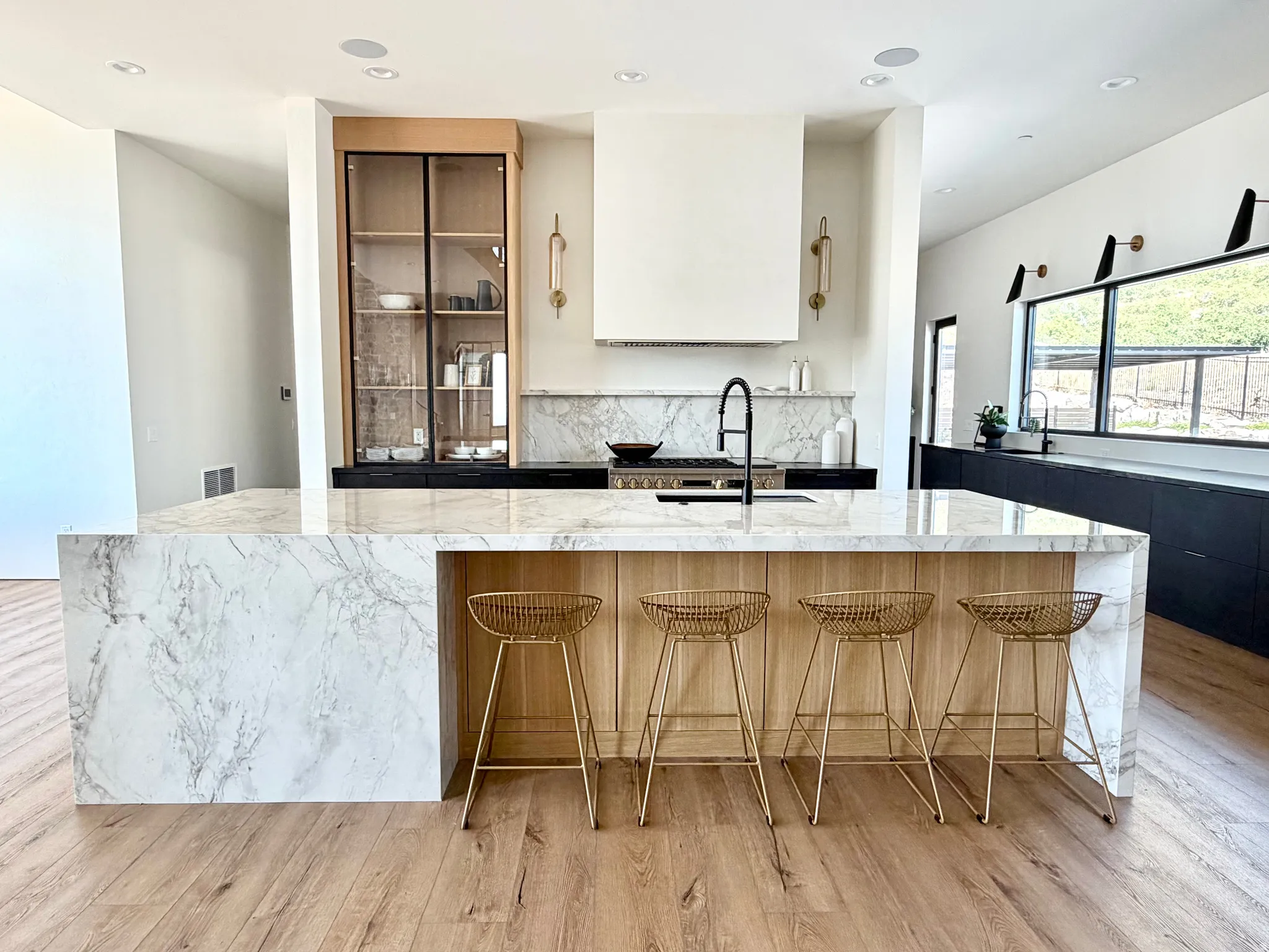 Waterfall island with stone finish, integrated cooktop, wood-finish flooring, built-in display cabinetry, and a full-height stone backsplash