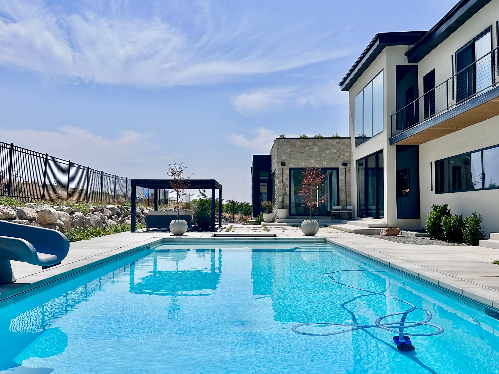 Outdoor swimming pool with integrated slide, contemporary multi-story exterior, stone-clad accent walls, large format windows, and a pergola-covered patio area