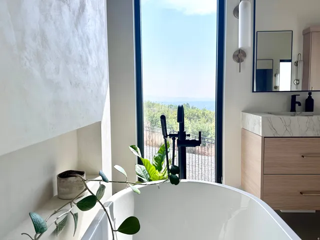 Freestanding soaking tub with a black floor-mounted faucet, large windows, wood-finish vanity with a stone countertop, and a contemporary wall mirror