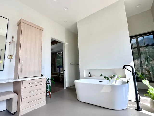 Freestanding soaking tub with matte black floor-mounted filler, built-in wall niche, vanity with wood-finish cabinetry, and a large window with black framing