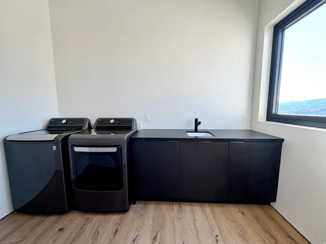 Laundry space featuring wood-finish flooring, a built-in sink with a matte black faucet, dark cabinetry, and an expansive window