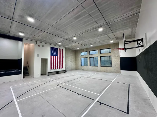 Indoor court featuring concrete flooring with court markings, exposed concrete ceiling with recessed lighting, multiple windows, and a wall-mounted basketball hoop