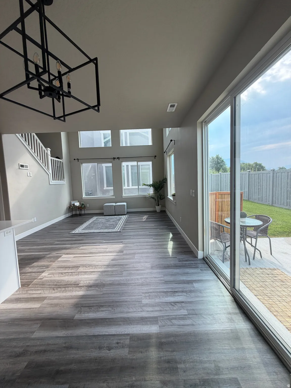 Expansive living area with wood-finish flooring, a contemporary chandelier, multiple large windows, and direct access to an outdoor patio with hardscaping