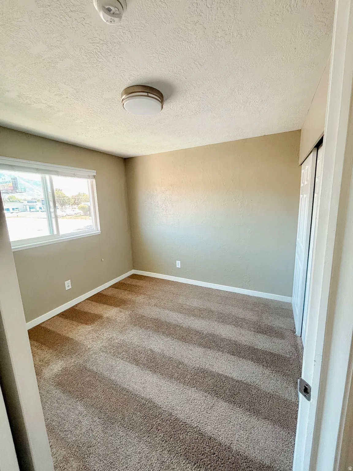 Wall-to-wall carpeting, neutral wall colors, flush-mount ceiling light, window with white trim, and bi-fold closet doors