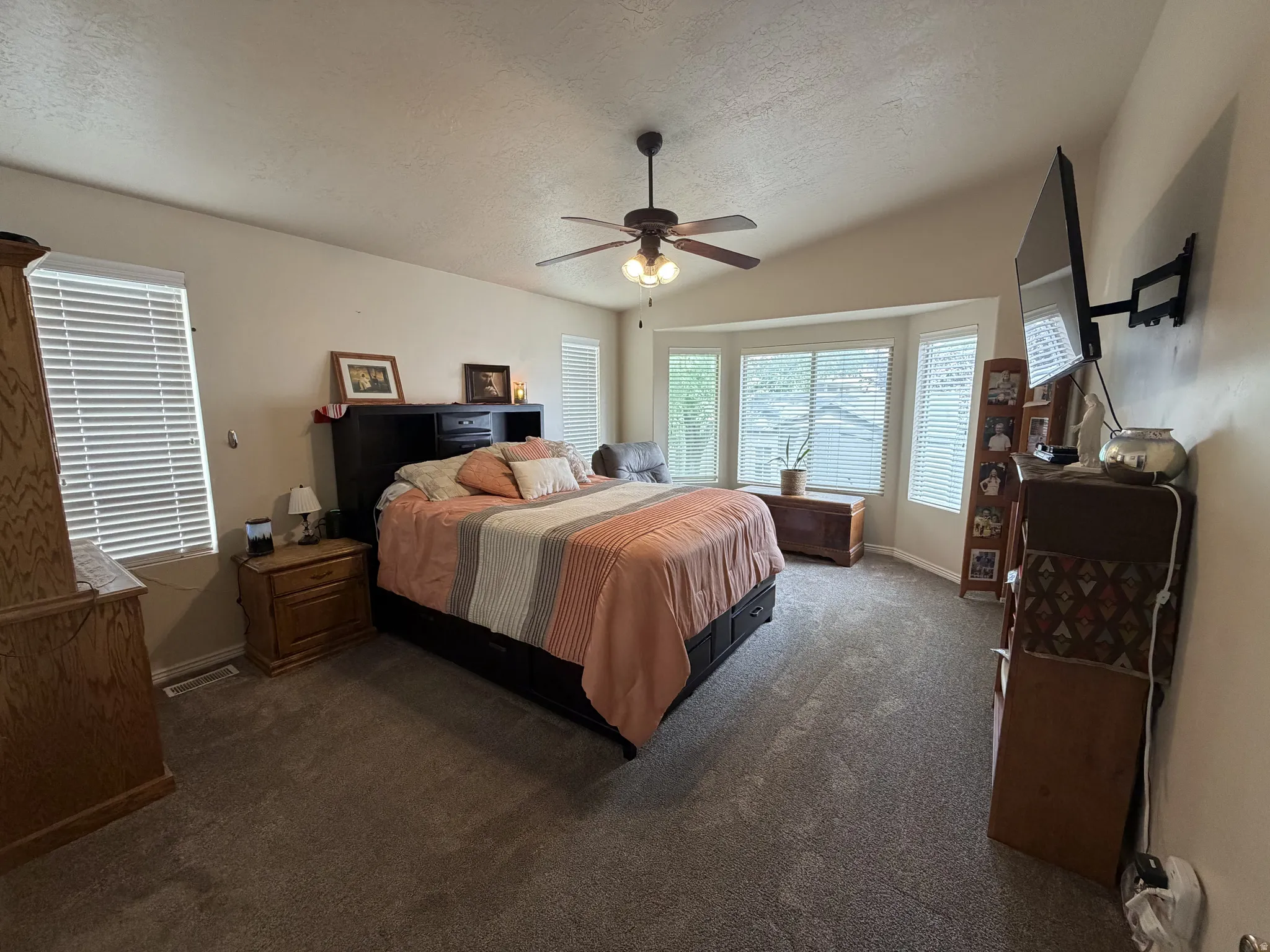 Primary suite 1. Spacious primary bedroom featuring abundant natural light from multiple windows, ceiling fan with integrated lighting, carpeted flooring, and wood-finish furnishings including dual nightstands and dresser storage