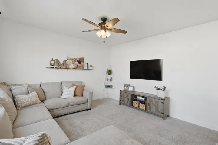 Vaulted ceiling, ceiling fan with integrated lighting, wall-mounted shelving, wood-finish media console, and neutral carpeting