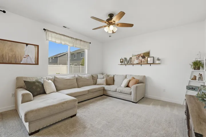 Carpeted flooring, white walls, large window, ceiling fan with light fixture, wall-mounted shelving