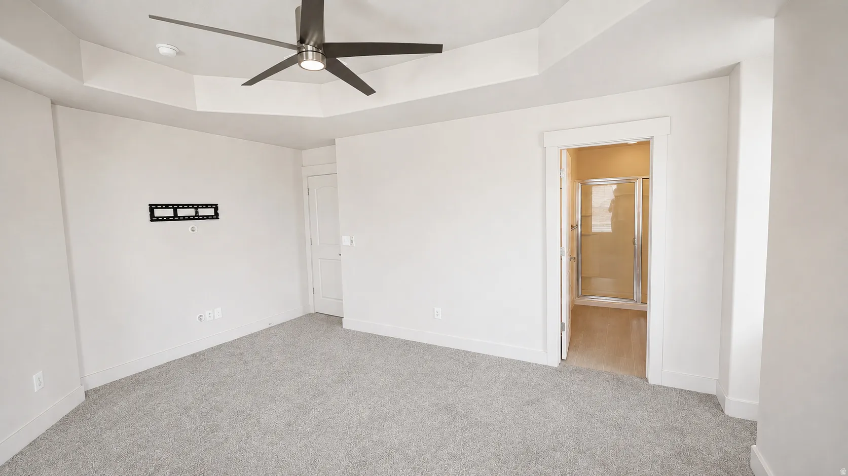 Tray ceiling, modern ceiling fan, wall-mounted television bracket, neutral carpeting, and a shower enclosure