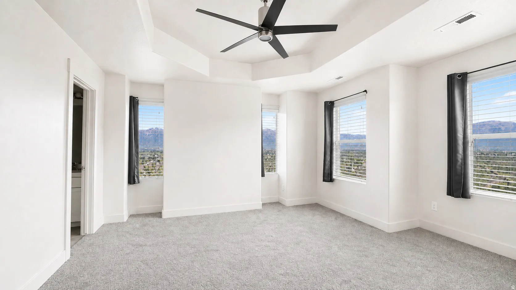 Tray ceiling with ceiling fan, multiple windows with blinds, carpeted flooring, and white painted walls