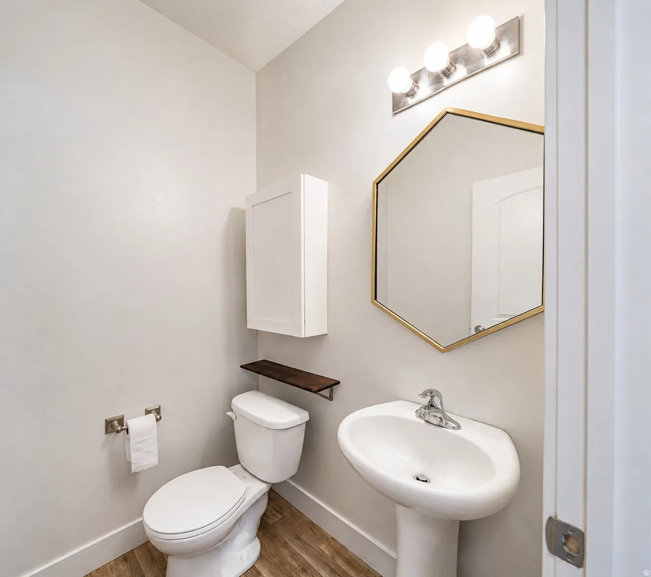 Pedestal sink with chrome faucet, hexagonal mirror with gold-tone frame, wall-mounted light fixture, wood-finish flooring, and white wall cabinet