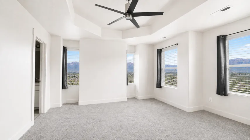 Tray ceiling with ceiling fan, multiple windows with blinds, carpeted flooring, and white painted walls