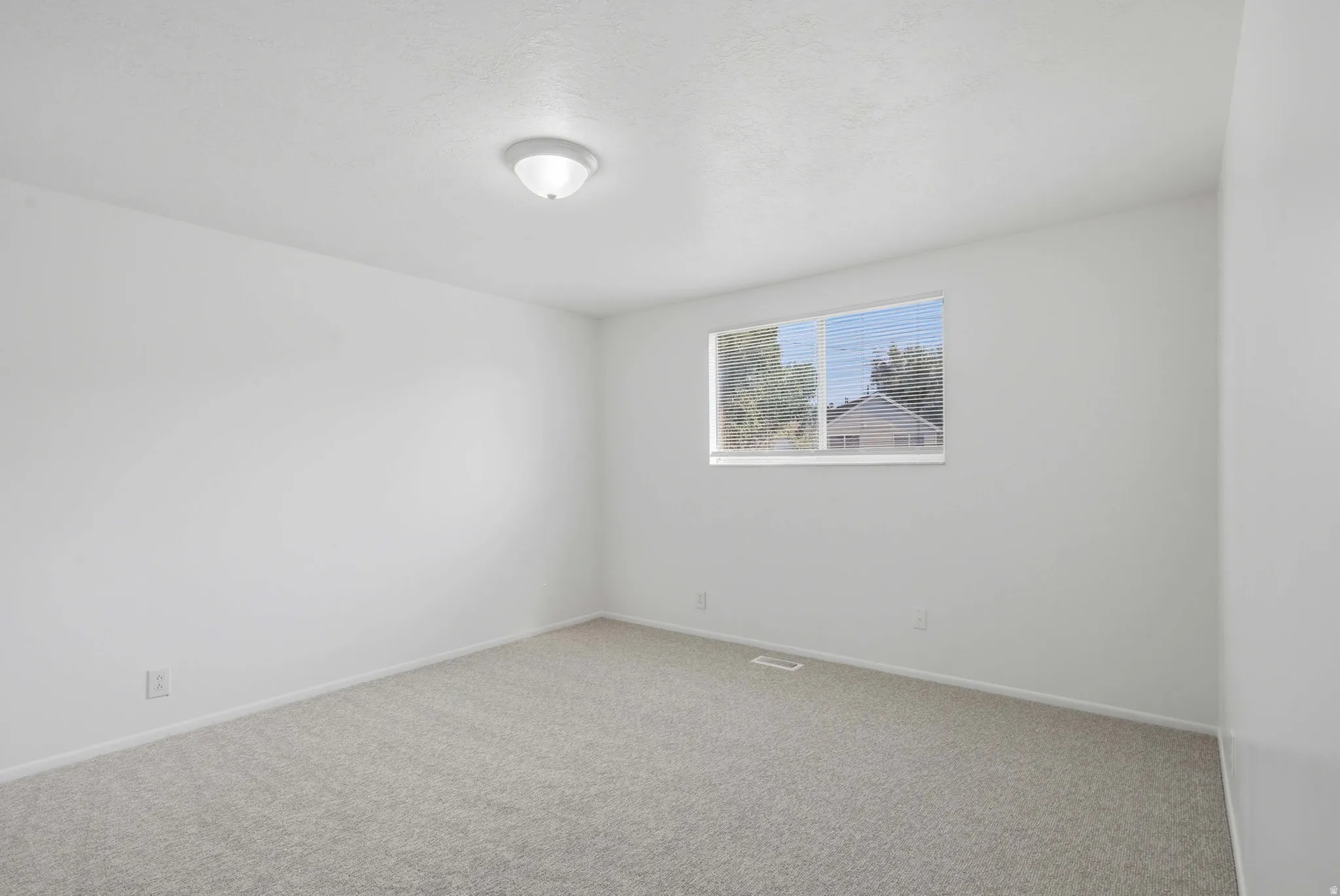 Spacious room featuring neutral carpeting, white walls, a single window with horizontal blinds, and a flush-mount ceiling light fixture