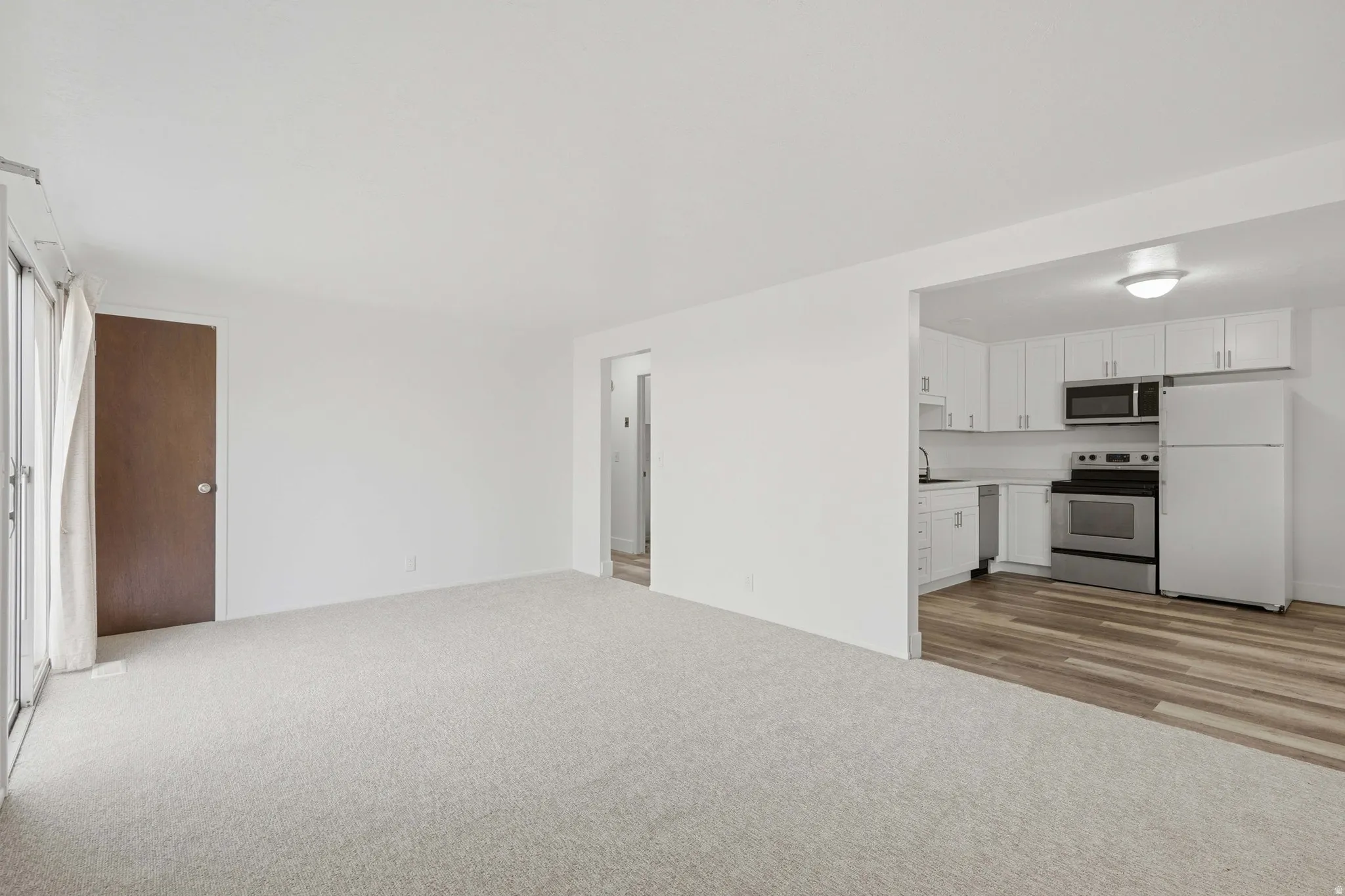 White kitchen cabinetry, stainless steel appliances, wood-finish flooring, light-toned carpet, and sliding glass door