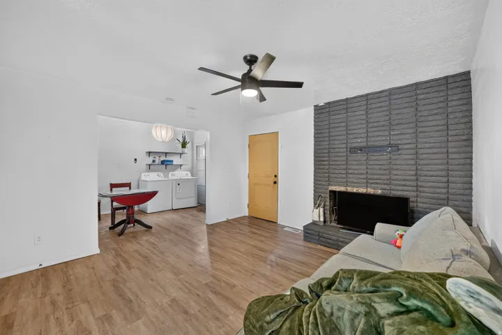Wood-finish flooring throughout, featuring a modern ceiling fan with lighting, a dark brick fireplace surround, and a dedicated laundry area with shelving