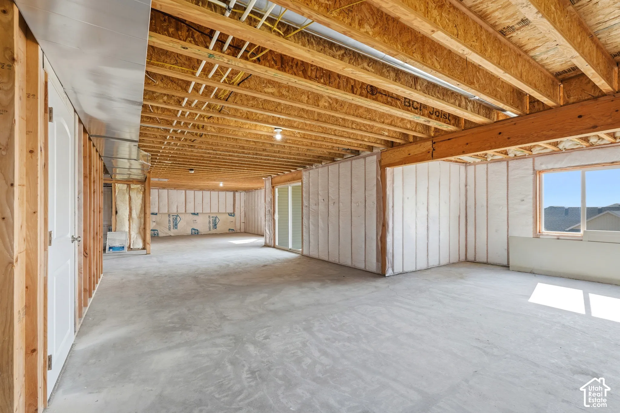 Unfinished basement with exposed wooden joists, concrete flooring, insulated walls, and a visible window