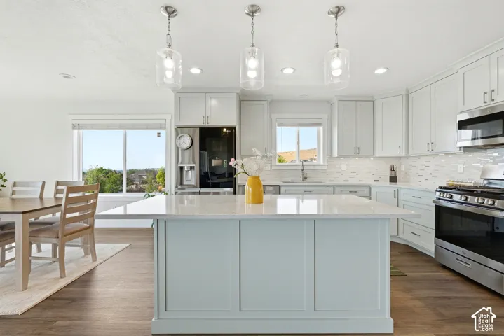 Central kitchen island with a light-toned countertop, light-colored cabinetry, stainless steel appliances, a tile backsplash, and wood-finish flooring
