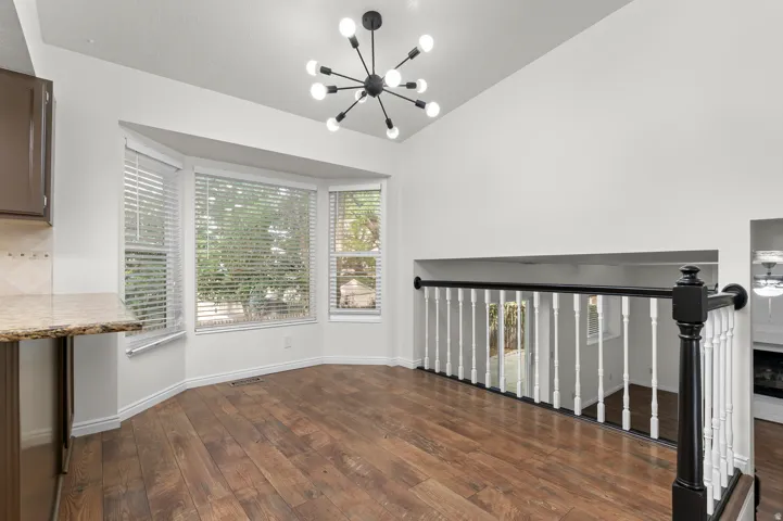 Vaulted ceiling with a modern sputnik chandelier, wood-finish flooring, bay window with blinds, and a staircase with a black and white banister