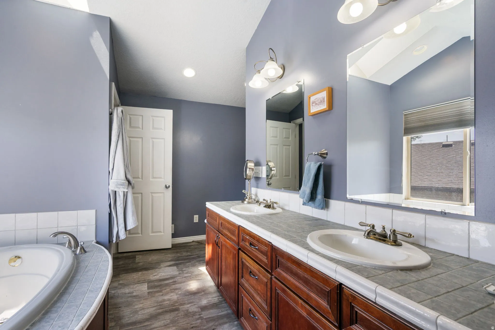 Vaulted ceiling bathroom featuring a built-in tub, dual sink vanity with wood-finish cabinetry, tile countertop, and wood-finish flooring