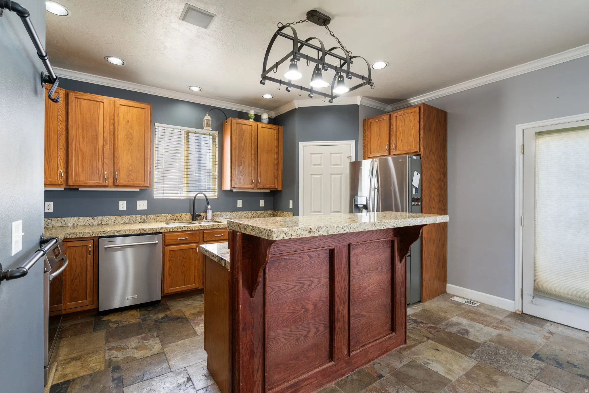 Kitchen featuring stone-look flooring, stainless steel appliances, wood cabinetry, granite countertops, and a central island with paneling