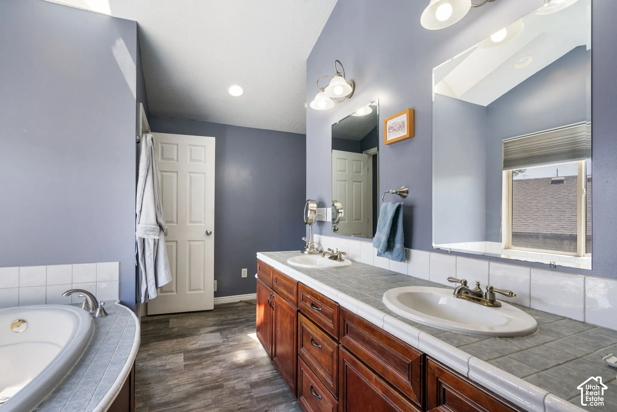 Vaulted ceiling bathroom featuring a built-in tub, dual sink vanity with wood-finish cabinetry, tile countertop, and wood-finish flooring
