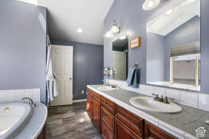 Vaulted ceiling bathroom featuring a built-in tub, dual sink vanity with wood-finish cabinetry, tile countertop, and wood-finish flooring