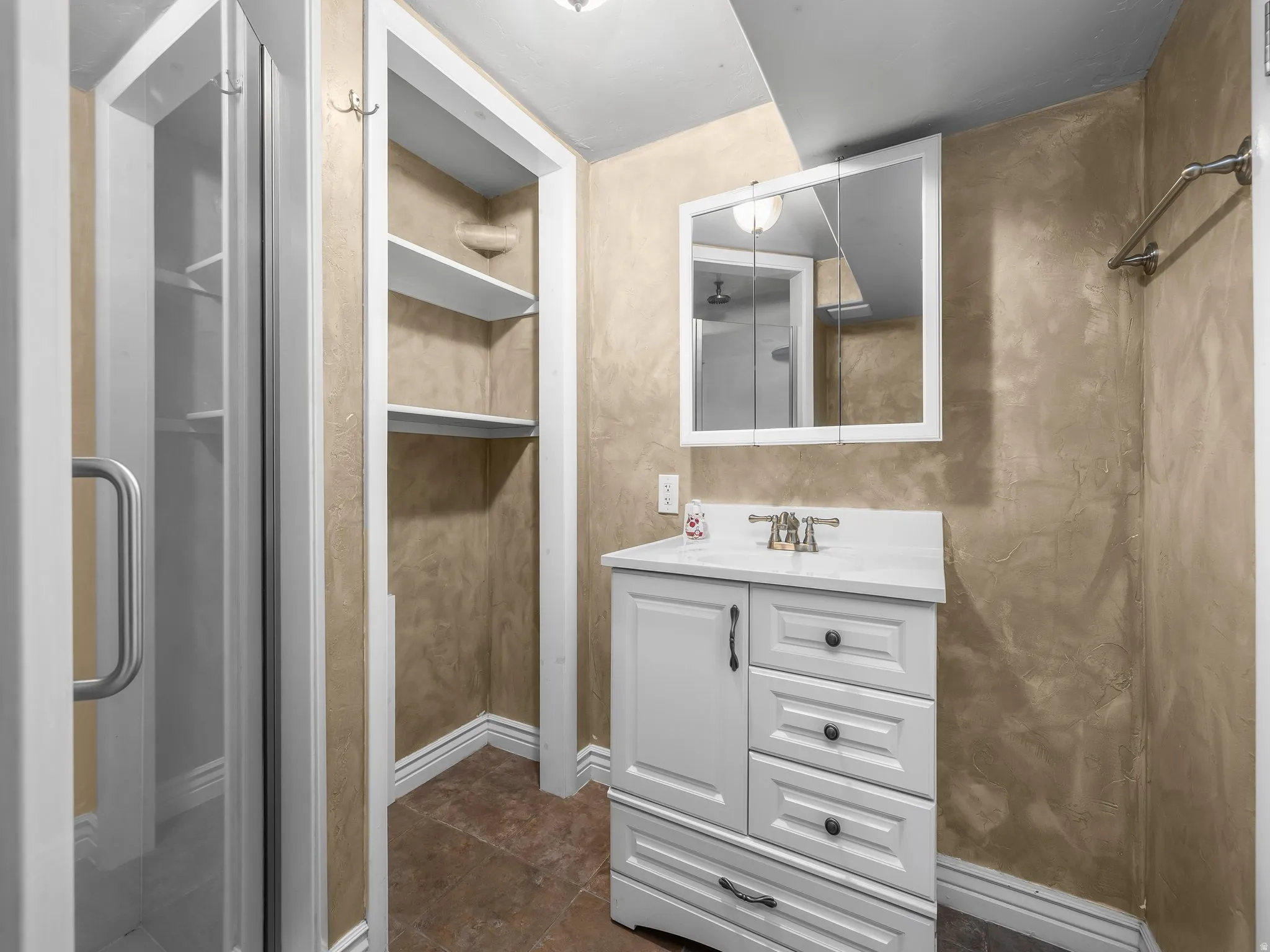 Basement bathroom. Glass-enclosed shower, built-in shelving, white vanity with integrated sink, mounted medicine cabinet, and towel bar