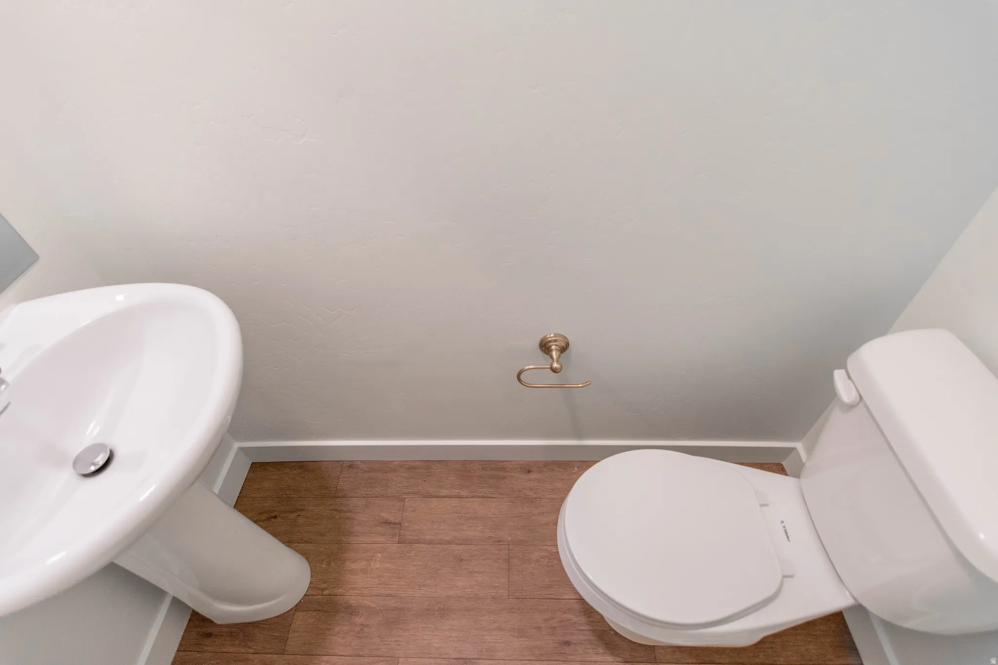 Powder room featuring a pedestal sink, white toilet, brass-finish toilet paper holder, and wood-finish flooring