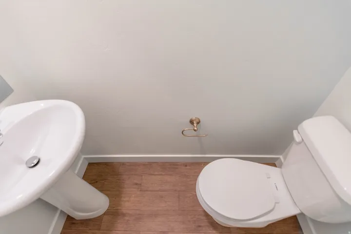 Powder room featuring a pedestal sink, white toilet, brass-finish toilet paper holder, and wood-finish flooring
