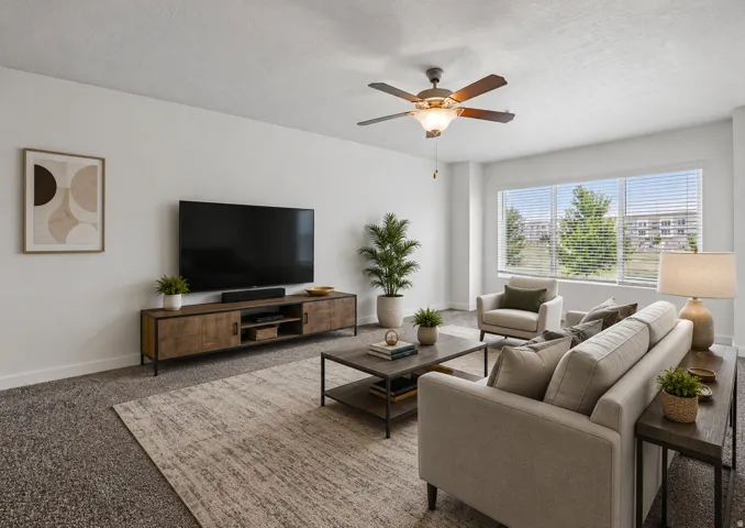 Spacious living area featuring a large window with blinds, a ceiling fan with integrated lighting, textured carpeting, and white baseboards