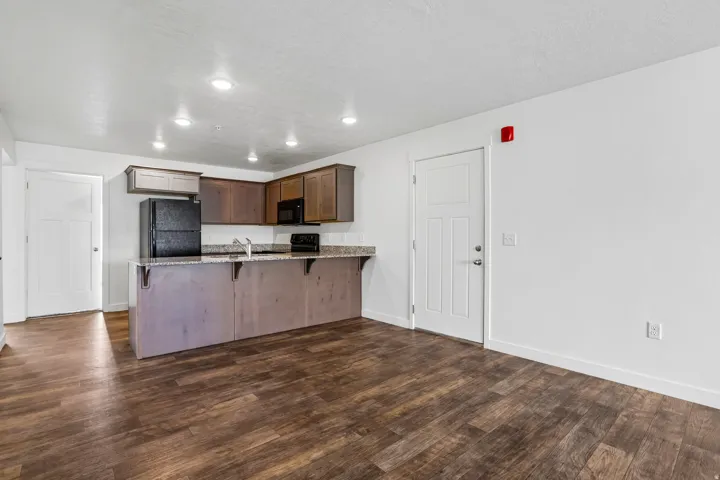 Open-concept living space with wood-finish flooring, a kitchen featuring dark wood cabinetry, black appliances, and a granite-style countertop, complemented by recessed lighting
