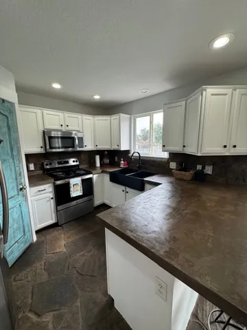 Kitchen featuring white shaker-style cabinetry, dark countertops, built-in stainless steel appliances, a black farmhouse sink, and flagstone flooring